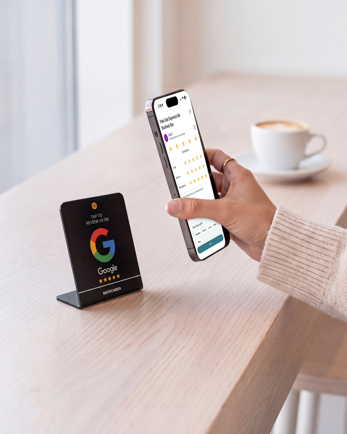 RATECARDS Google Review Stand Premium Black on a pale-oak coffee bar, customer holding her phone on the Google review screen