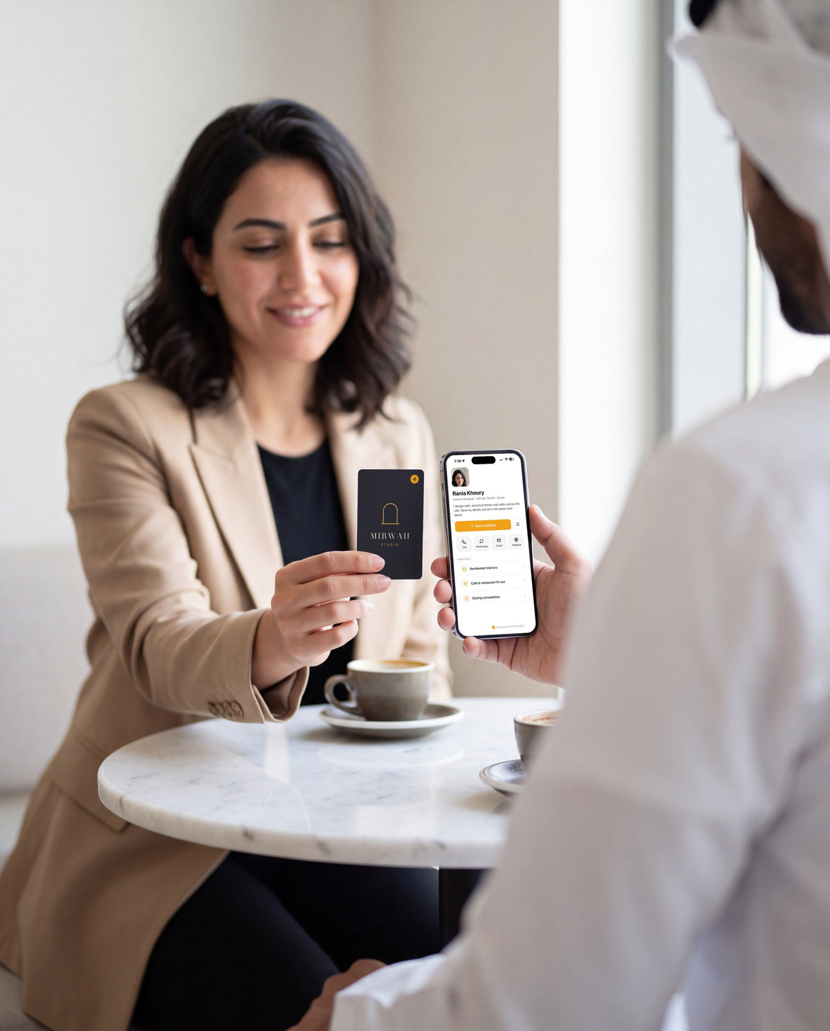 Two professionals at a café table: she holds out her RATECARDS NFC Business Card, his phone shows her contact page